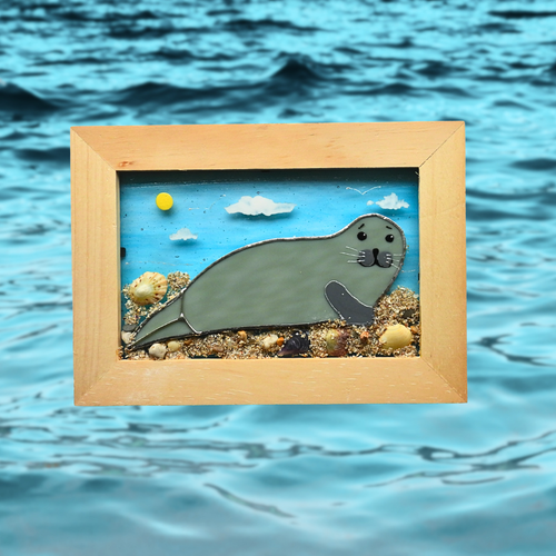 Framed glass artwork of a seal on a beach with ocean waves in the background