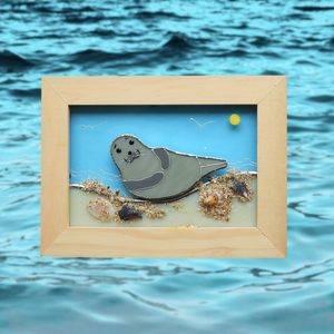 Framed glass artwork of a seal on a beach with ocean waves in the background
