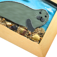 Load image into Gallery viewer, Framed glass art of a seal on a beach with sand and shells stuck on
